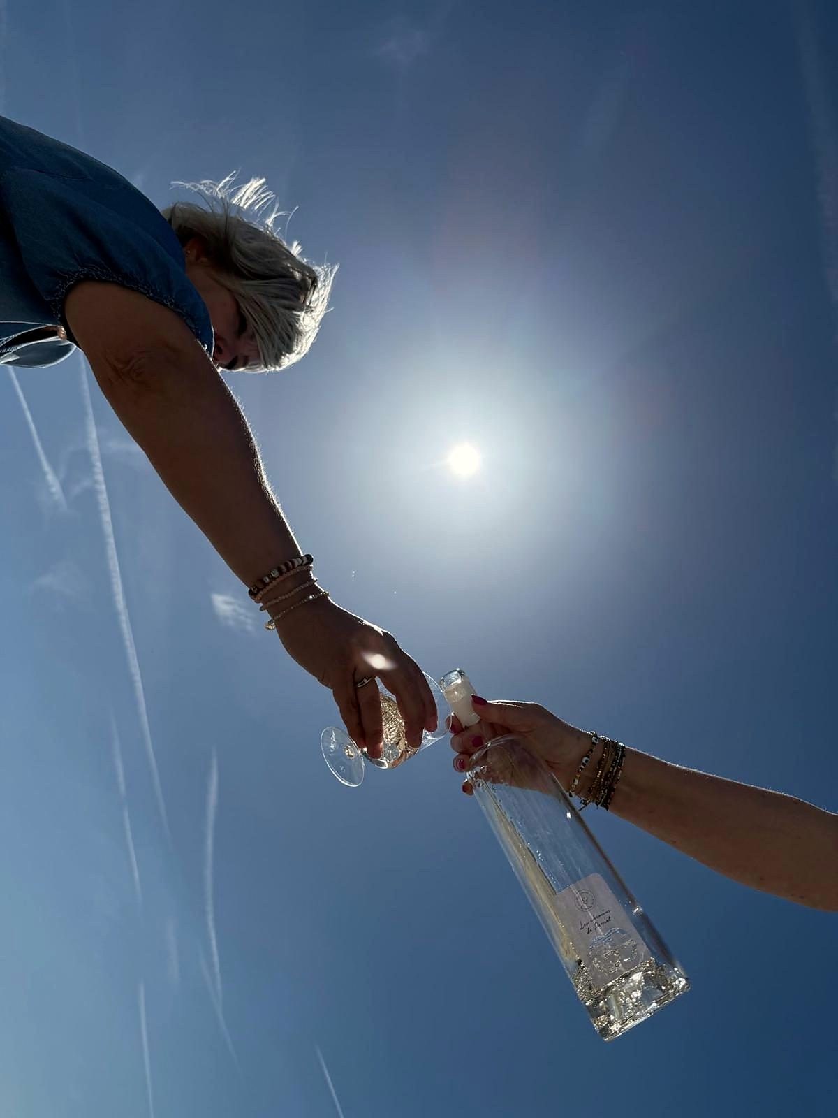 Twee handen die een glas en een fles naar elkaar brengen, tegen de zon